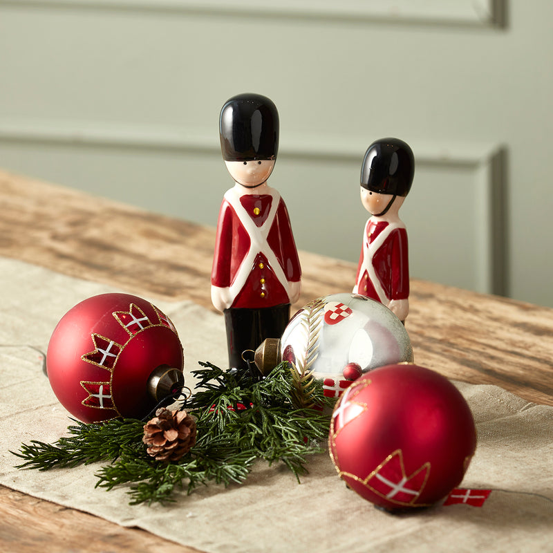 Røde julekugler med Dannebrog på et bord sammen med garderfigurer, grangrene, en kogle og en sølvfarvet julekugle.