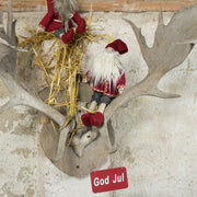 Rødt ‘God Jul’-skilt hængende under et dekorativt gevir med to røde nisser og halm på en rustik væg.
